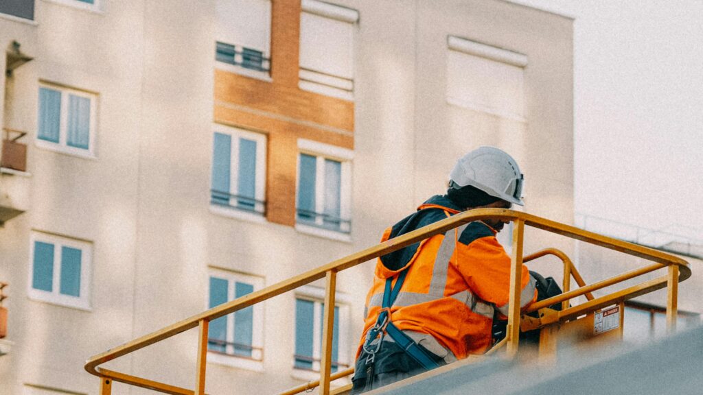 A construction worker in safety gear operates a lift near an apartment building.